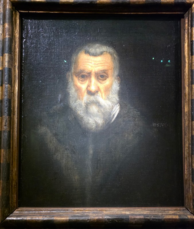 Portrait d'un homme avec une barbe grise et des cheveux clairs, portant des vêtements sombres, sur un fond sombre.